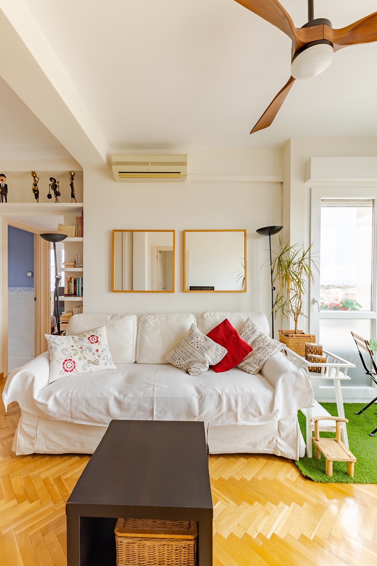 A bright living area features a white sofa adorned with patterned cushions and a bold red accent pillow. Two rectangular mirrors reflect light and space, while a small coffee table sits in front. Lush greenery and natural light enhance the inviting atmosphere.