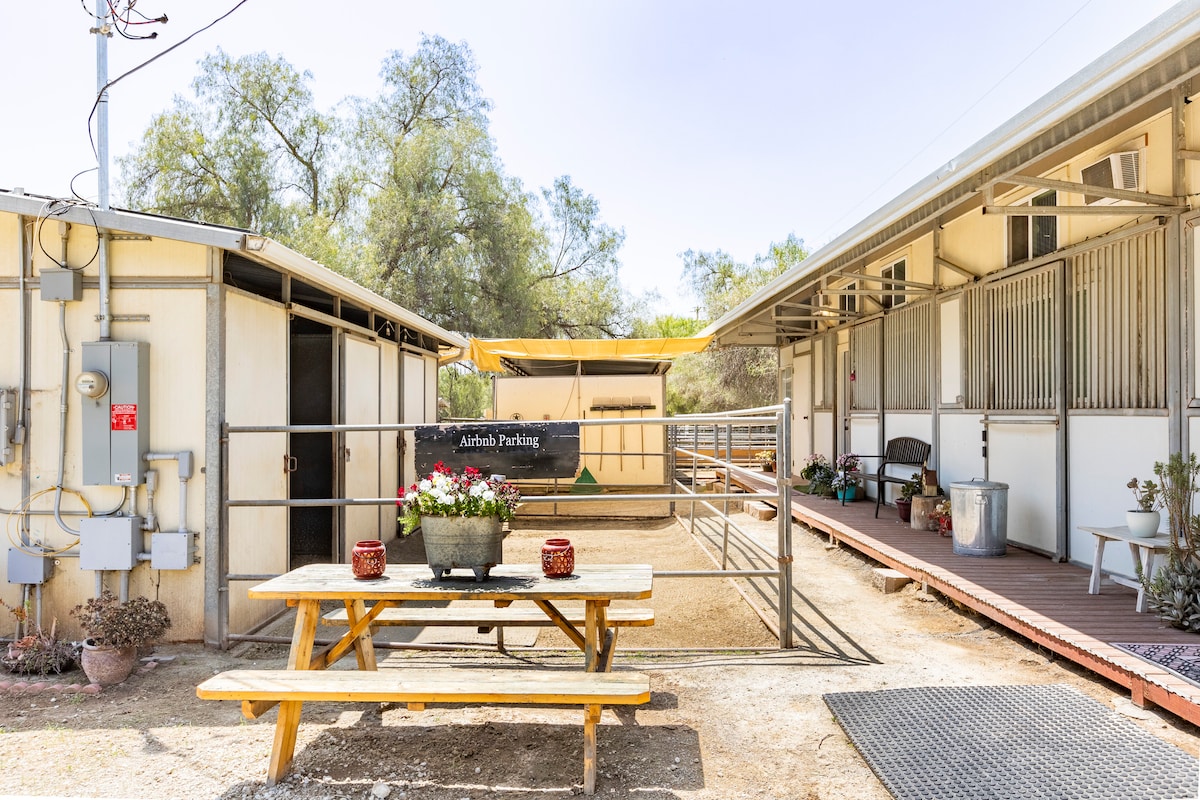 A picnic table with decorative pots is positioned outside, surrounded by a fenced area. Two structures display horse stalls, with shaded outdoor spaces visible. Natural greenery frames the scene, adding to the rustic charm of the ranch.
