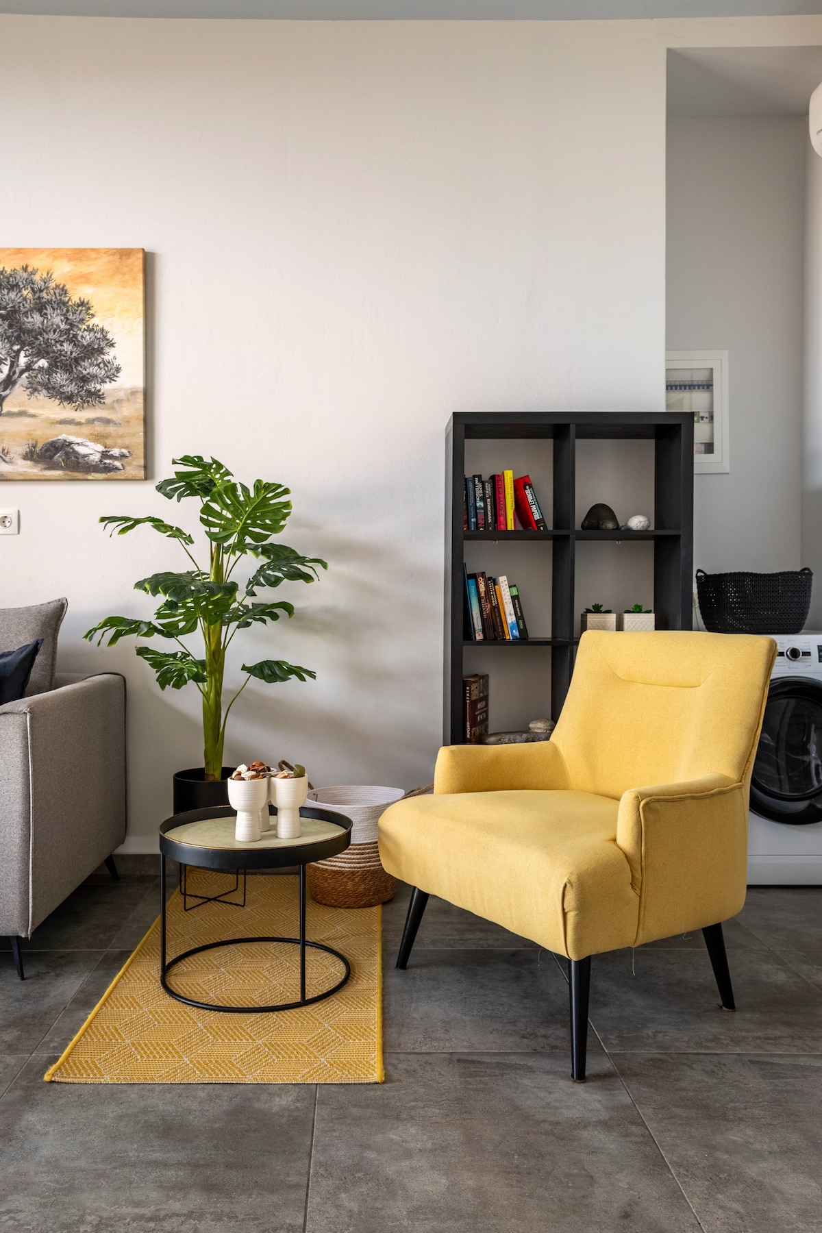 A bright corner of the living area features a yellow armchair beside a small round table with two cups. A bookshelf with decorative items and books stands nearby, while a potted plant adds a touch of greenery to the space.
