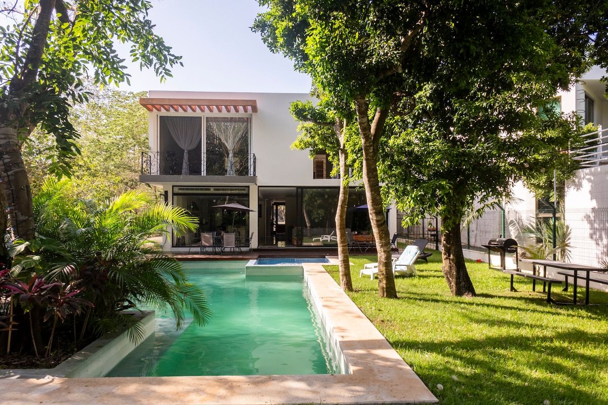 A spacious backyard features a serene swimming pool surrounded by lush greenery and palm trees. Sun loungers are positioned beside the pool, with a dining table visible in the area. The two-story house, with large windows, reflects natural light and an inviting ambiance.