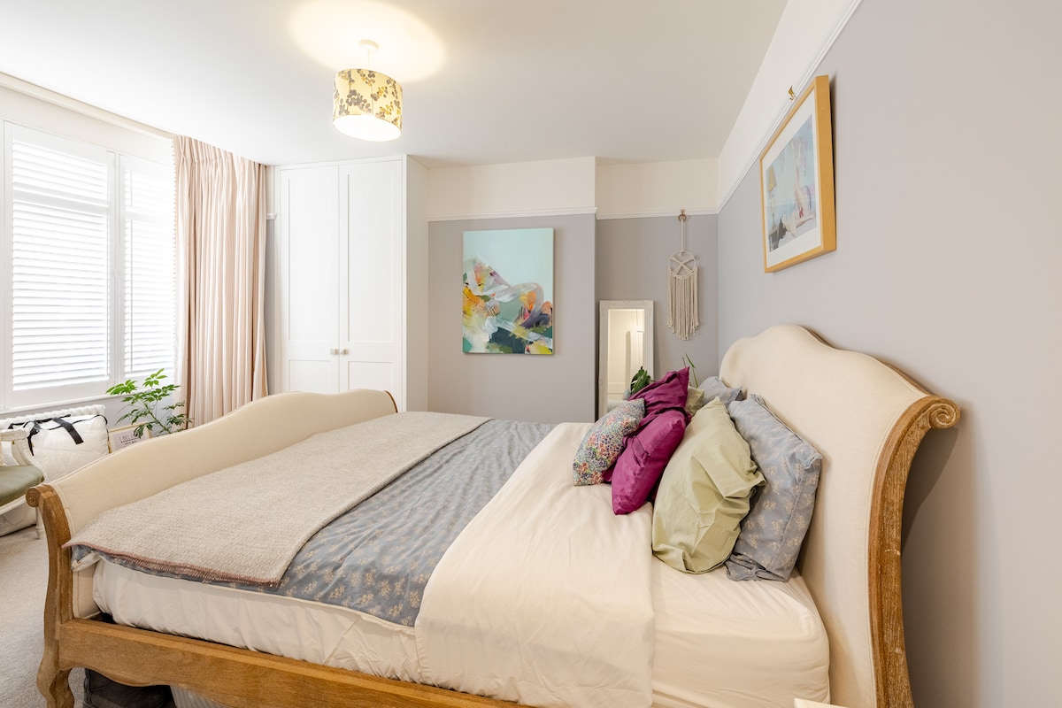 A spacious bedroom features a large double bed with a mix of colorful pillows against a pale grey wall. A piece of abstract artwork hangs above the bed, and natural light floods in through window treatments. A stylish light fixture complements the warm tones of the room.