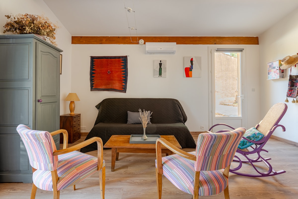 A cozy living area is presented with a brown sofa and a wooden coffee table, complemented by two striped armchairs. A vibrant wall hanging adds color, while a door leads outside. A rocking chair is also featured, enhancing the relaxed atmosphere.