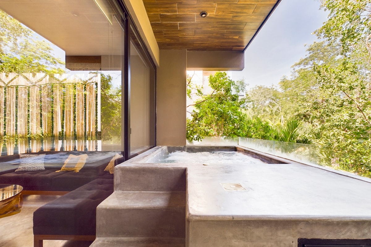 A private terrace features a small plunge pool, complemented by a concrete surround. Large glass doors offer views of the lush jungle landscape, while a cozy seating area with dark cushions is arranged beside the pool, creating a serene outdoor space.