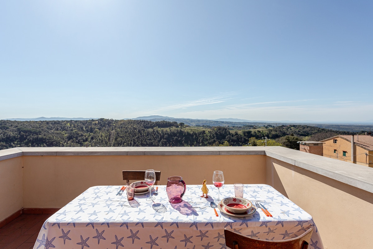 An outdoor dining table is set with tableware, positioned on a spacious terrace. The view extends over lush green hills towards the horizon, where a clear blue sky meets distant mountains. Sunlight brightens the scene, creating a serene atmosphere.