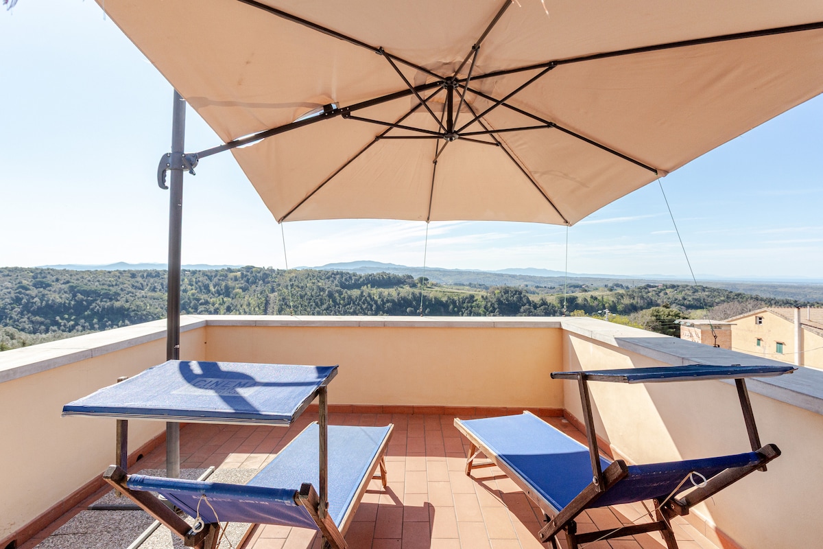A spacious terrace is furnished with two lounge chairs under a large umbrella, providing shade. The expansive view of rolling hills and trees is visible in the background, creating a serene outdoor space for relaxation.