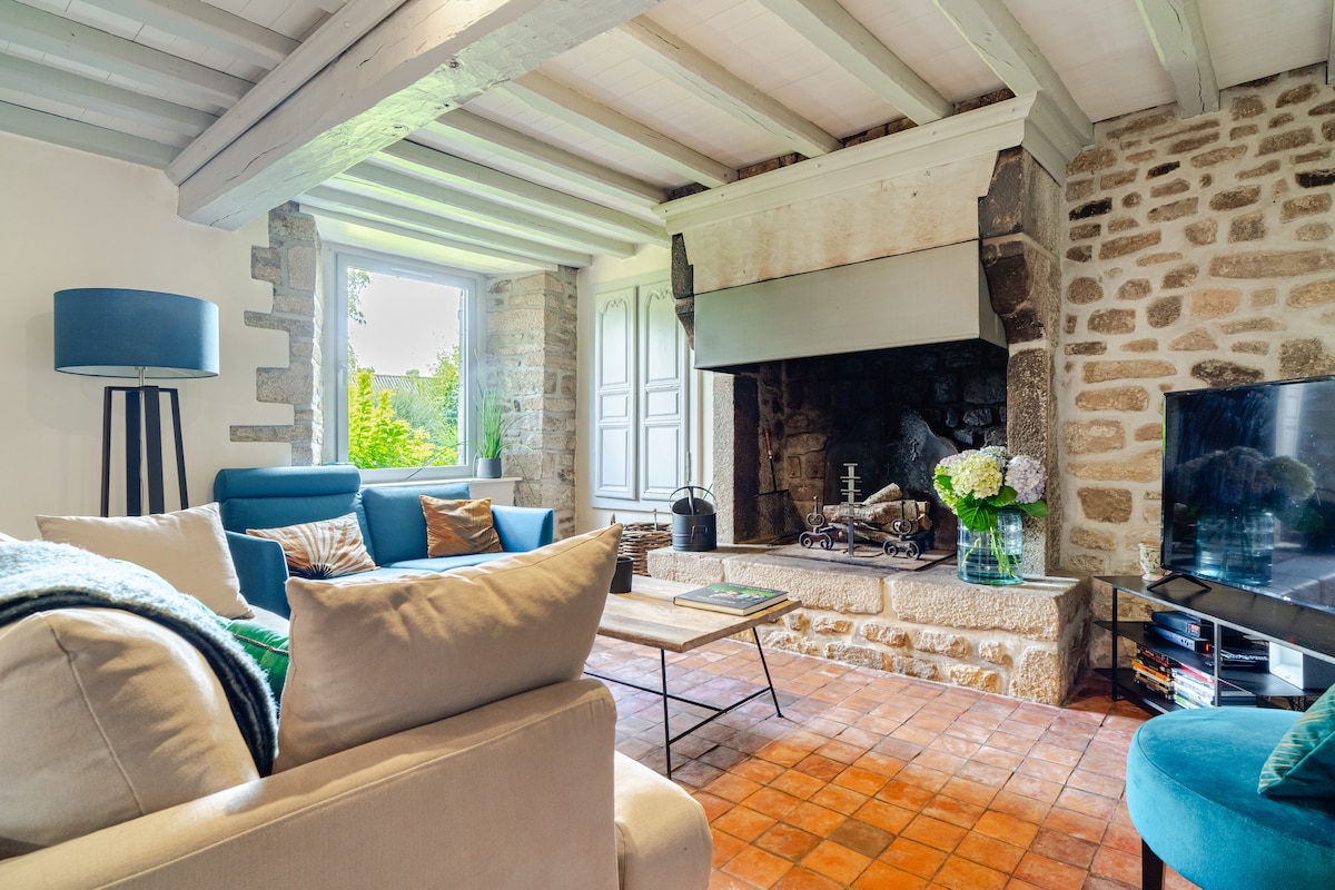 A living area features stone walls and a rustic fireplace, enhancing the cozy ambiance. A casual seating arrangement includes a blue sofa and a neutral-toned couch, while a coffee table sits in front. Natural light enters through a window, highlighting the room's inviting decor.