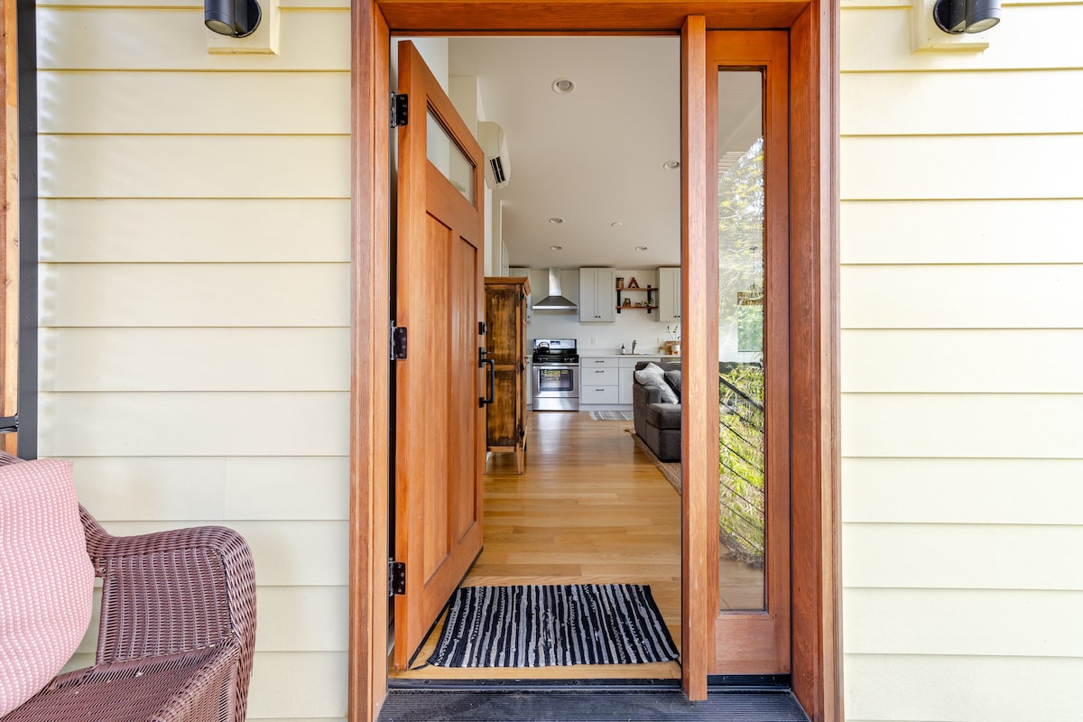 An inviting entryway features a solid wood door flanked by large windows, allowing natural light to illuminate the space. A patterned rug is placed on the threshold, leading into a bright open-plan living area that includes a kitchen and seating area.