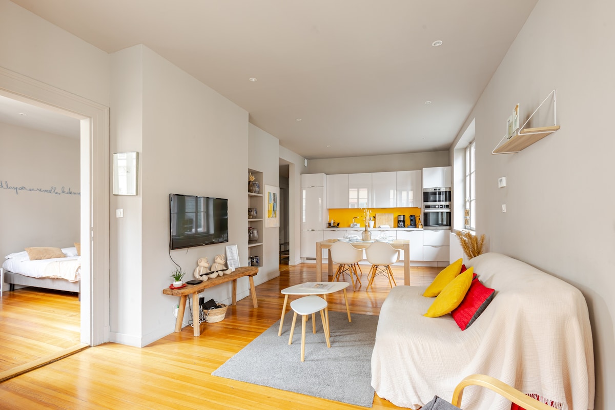 The spacious living area features hardwood floors, natural light from large windows, and modern furnishings. A white sofa with colorful cushions is positioned near a flat-screen TV. A dining table with chairs is set beside the kitchen, which is brightened by yellow backsplash tiles.