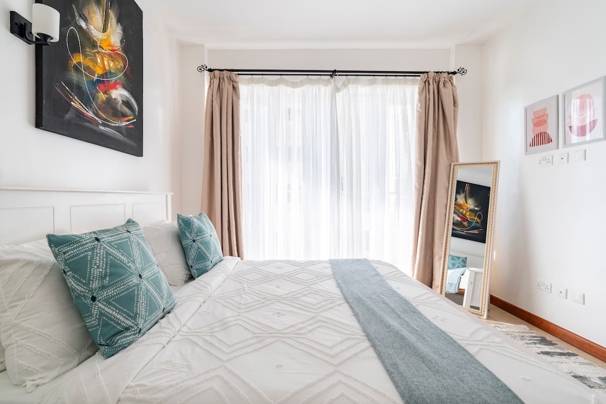 חדר שינה מודרני בקילימאני עם מיטת קינג סייז, אמנות מופשטת על הקיר, מראה באורך מלא, חלון גדול עם אור טבעי, עיצוב גיאומטרי ושטיח מעוצב. אידיאלי לנסיעות עסקים או לשהיות ממושכות ליד ווסטלנדס ו-JKIA.