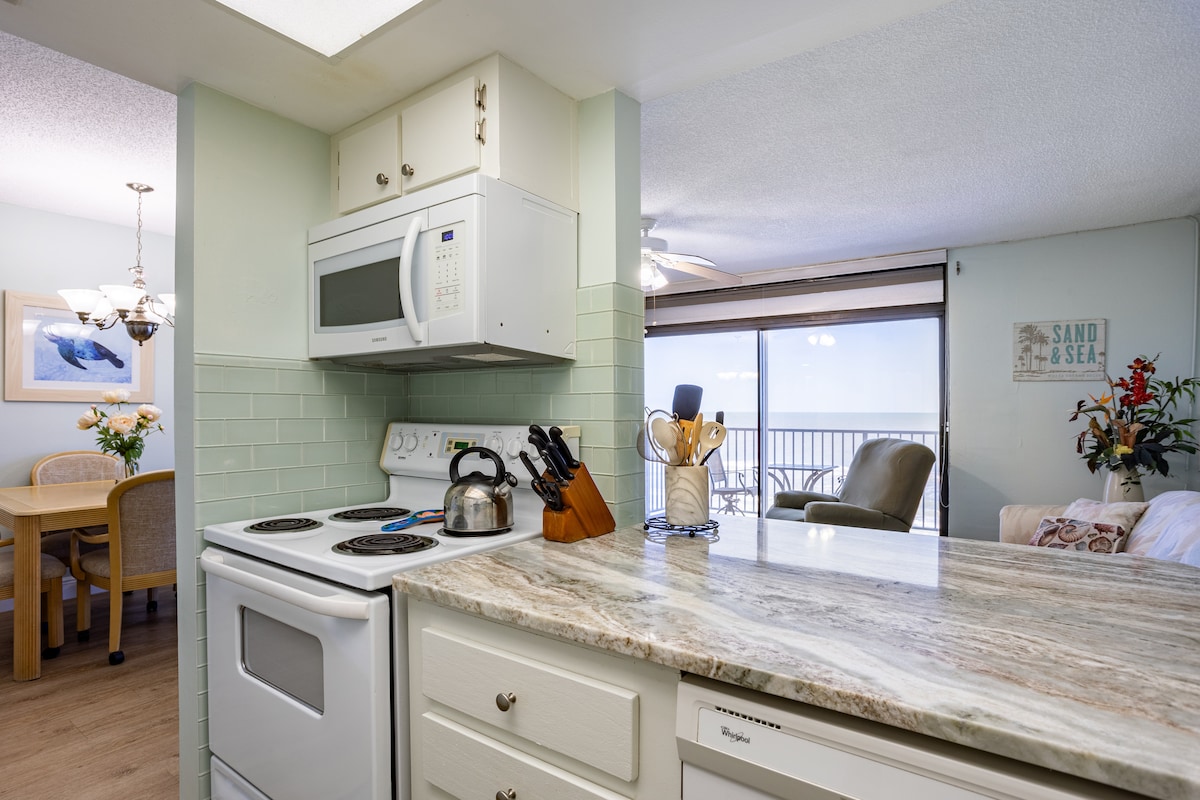 A well-equipped kitchen features a white stove and microwave, complemented by a sleek countertop. Various kitchen utensils are neatly arranged, and a dining area with a table and chairs is visible in the background. Natural light floods the space, providing a warm atmosphere.