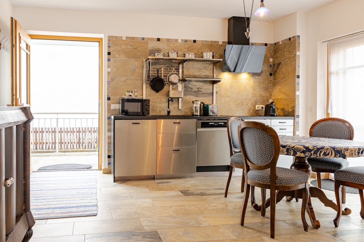 The kitchen space features modern appliances, including a refrigerator, microwave, and dishwasher, integrated into a stylish layout. A wooden dining table with elegant chairs is situated nearby, while the entrance leads to a balcony, allowing natural light to brighten the area.