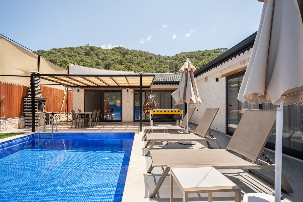 A private outdoor pool area features a clear blue swimming pool surrounded by several reclining lounge chairs. A shaded pergola provides outdoor seating, while an umbrella stands nearby. Lush green hills create a serene backdrop, complementing the relaxing atmosphere.