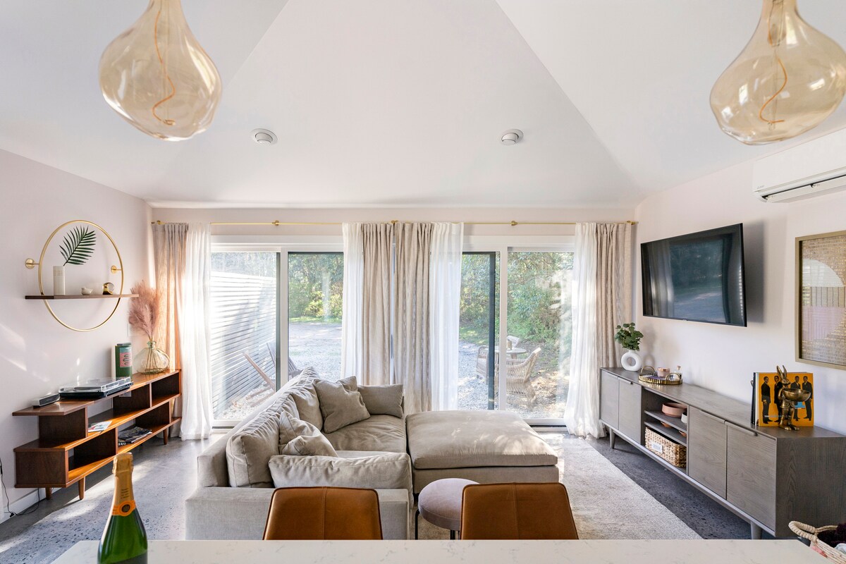 A bright living space features large sliding glass doors that provide natural light and a view of the outdoors. A plush sectional sofa is positioned in the center, complemented by a modern entertainment unit and decorative accents on the walls. Elegant pendant lights hang from the ceiling.