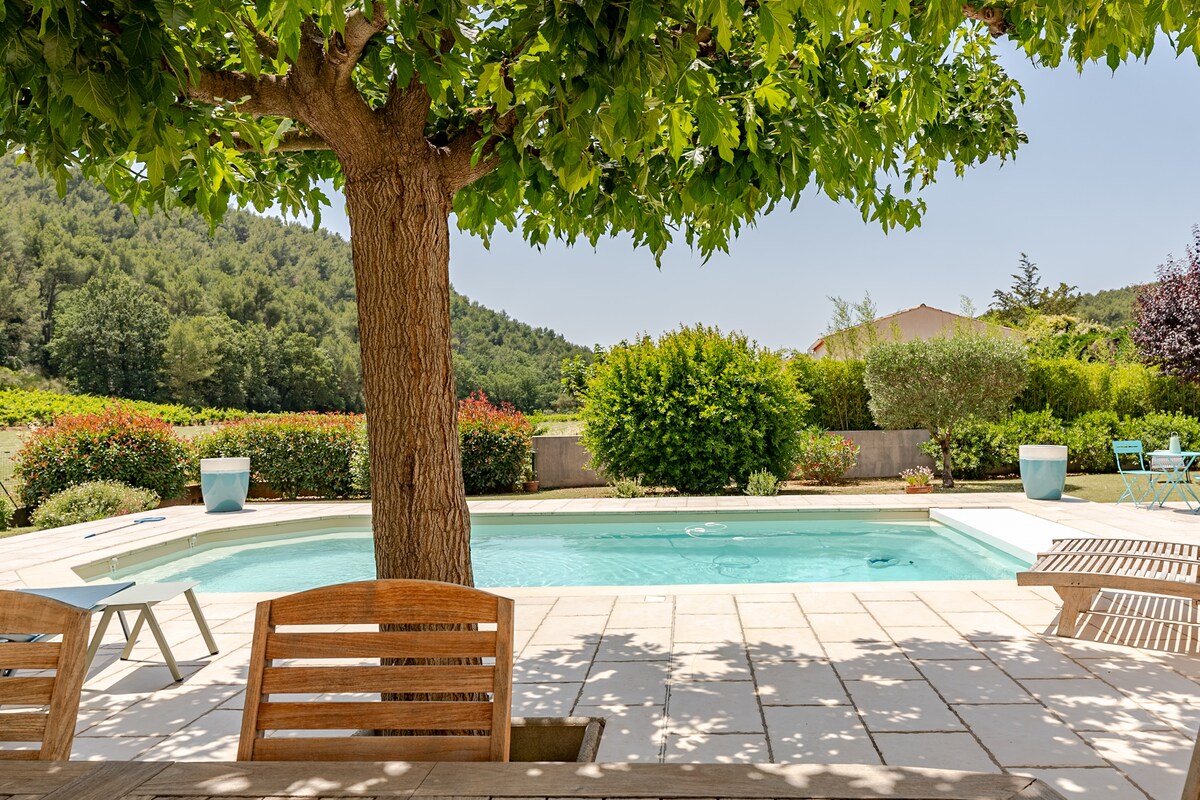 A serene outdoor area features a well-maintained swimming pool surrounded by lush greenery and flowering bushes. A large tree provides shade, while a wooden table and chairs are arranged nearby for relaxation. The setting offers a peaceful ambiance, ideal for unwinding in nature.