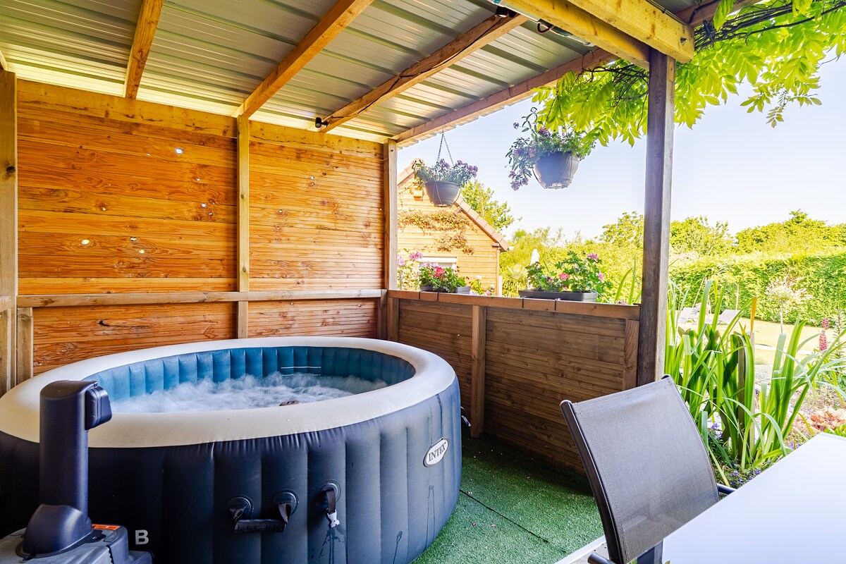 An outdoor covered area features a spacious inflatable jacuzzi surrounded by wooden paneling, with potted plants hanging above. A table and chair set is visible nearby, complemented by a green landscape beyond, creating a tranquil setting for relaxation.