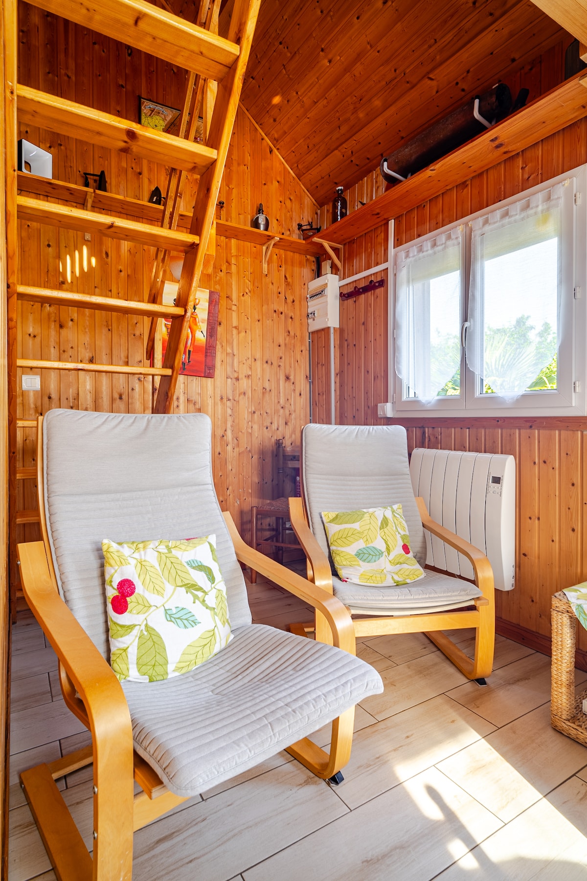 Two comfortable armchairs with cushions are positioned in a wooden interior, featuring warm tones. Natural light enters through a window, enhancing the cozy atmosphere. A wooden ladder leads to the mezzanine, while additional decorative elements can be seen on the walls.