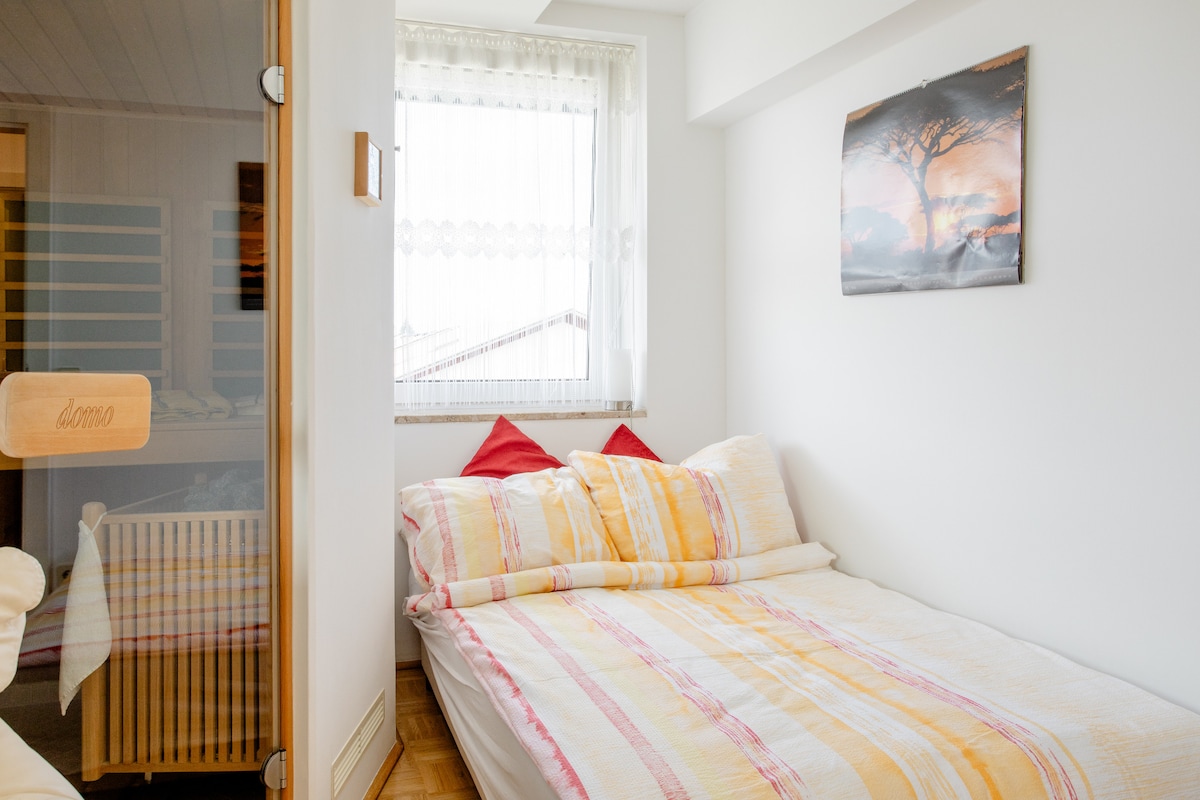 A bright sleeping area features a spacious bed with light-colored linens, accented by two red decorative pillows. Natural light filters through a window dressed with sheer curtains, while a piece of artwork depicting a tree is displayed on the adjacent wall.