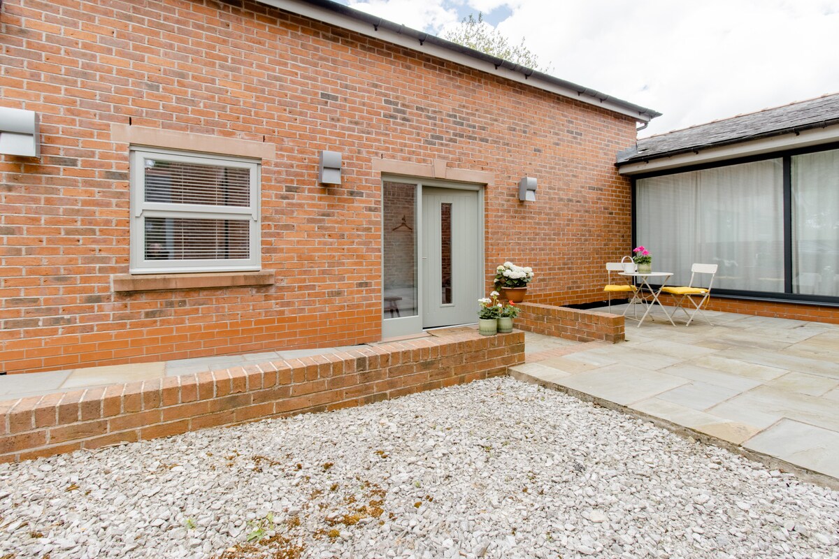 The exterior of the annexe showcases a modern brick facade with a grey door flanked by two small windows. A stone patio area is seen to the right, featuring a small bistro table and chairs. Flower pots add a touch of greenery to the entrance.