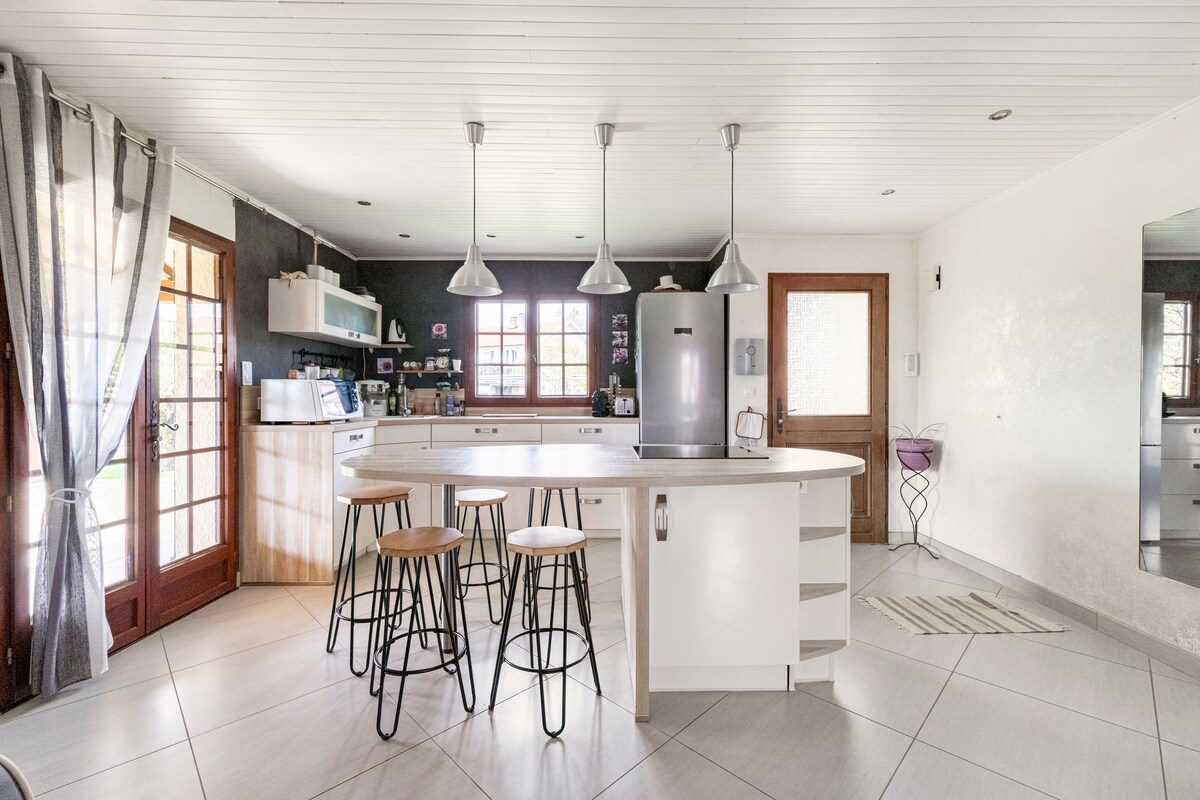 A spacious kitchen area features a central island with three tall stools. Modern stainless steel appliances and cabinetry provide ample storage. Natural light enters through large windows, complemented by elegant gray curtains. The open layout connects to the dining and living spaces, enhancing a sense of openness.