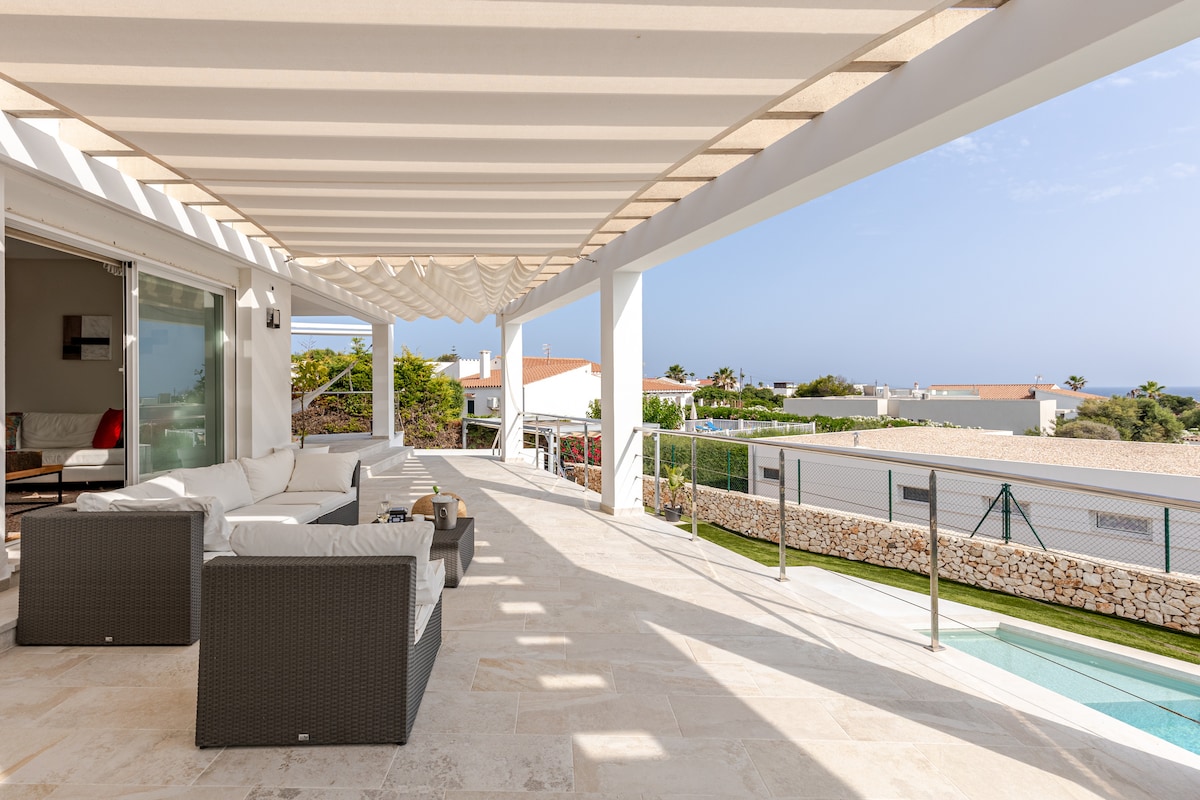 An expansive terrace is shown, featuring a lounge area with comfortable seating arrangements. The area is shaded by a slatted roof, while a glimpse of the swimming pool and landscaped surroundings can be seen in the background, allowing for a relaxing outdoor experience.