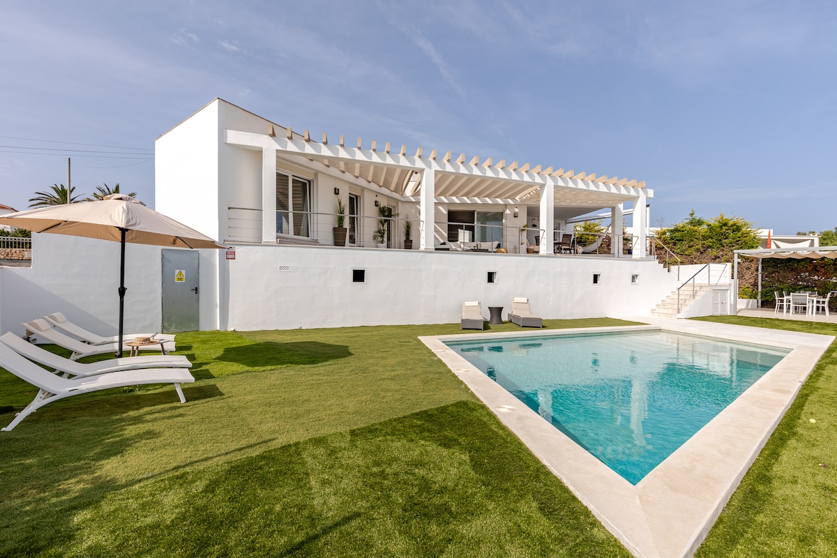 A modern villa is depicted, featuring a spacious outdoor area with a lush green lawn. A clear swimming pool reflects the sky, accompanied by reclining lounge chairs. The white building above offers shaded seating areas, providing an inviting space for relaxation.
