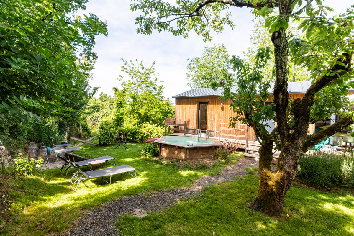 A tranquil outdoor setting features a wooden structure surrounded by greenery. Sun loungers are placed on the grass, and a hot tub is positioned nearby. A winding path leads through the area, with trees providing shade and enhancing the natural ambiance.