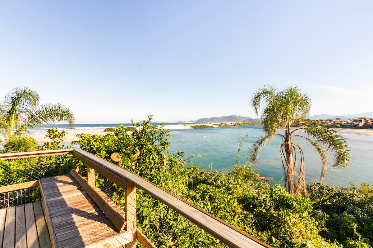 A scenic view of the coastline is framed by lush greenery and palm trees. The water glistens in the sunlight, and distant mountains are visible on the horizon. A wooden railing is positioned in the foreground, enhancing the natural beauty of the landscape.