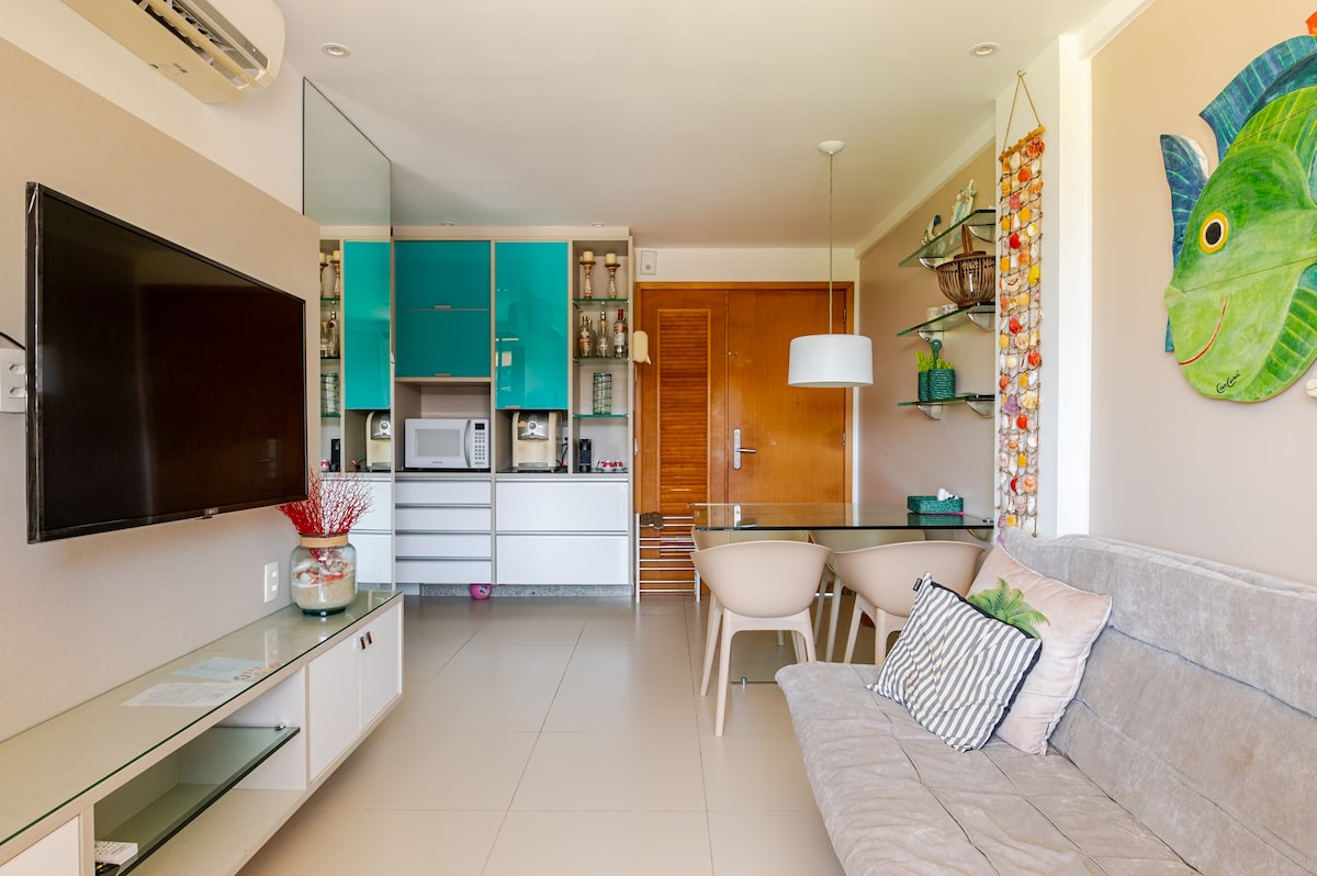 An open living area features a light gray sofa and a glass dining table surrounded by four chairs. A large television is mounted on the wall, with turquoise cabinetry displaying decorative items and appliances. Natural light brightens the space through a nearby window.
