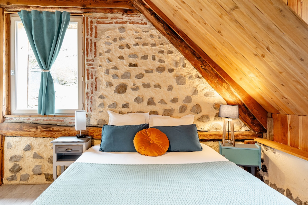 A cozy sleeping area is presented with a spacious bed adorned with teal and white bedding. A large orange pillow adds a pop of color. Nearby, a wooden lamp provides soft lighting next to a nightstand. Natural light enters through a window draped with light blue curtains against a rustic stone wall.