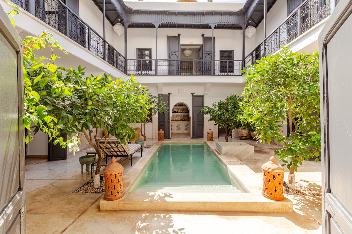 A serene courtyard is highlighted by a tranquil swimming pool surrounded by lush greenery and decorative orange lanterns. Elegant balconies overlook the space, offering a peaceful retreat. The smooth stone flooring complements the natural elements, creating a harmonious ambiance.