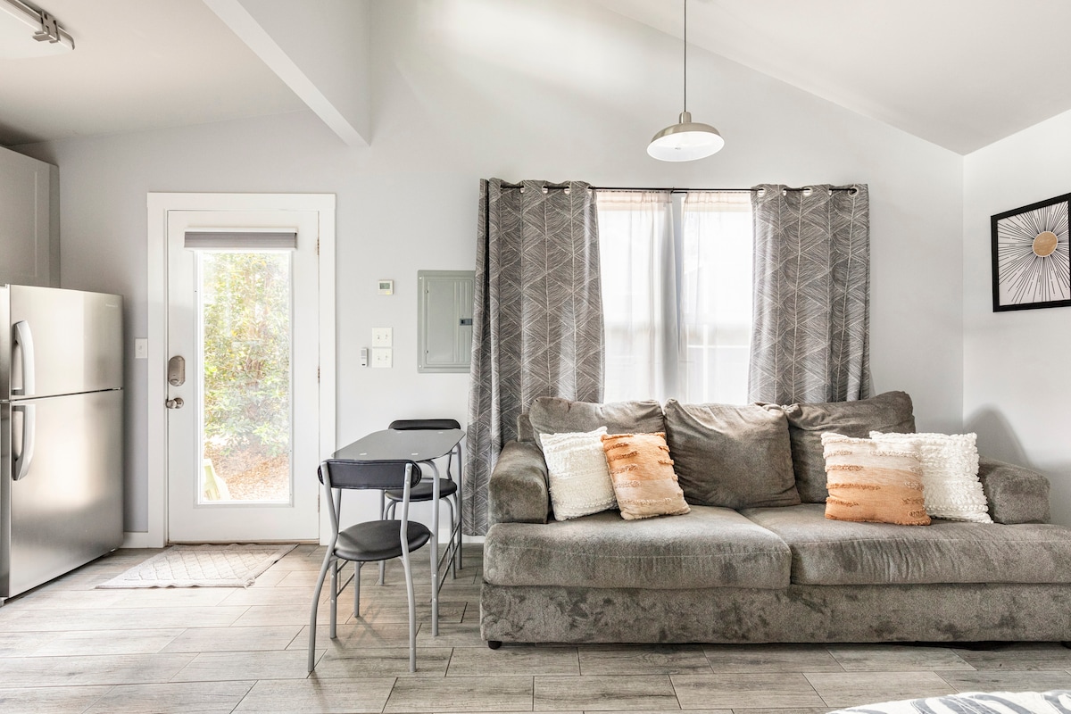 A modern living area features a cozy couch adorned with decorative pillows. A small dining table and chairs are positioned nearby. Natural light enters through a window dressed with light curtains, and a door leads outside, enhancing the welcoming space.