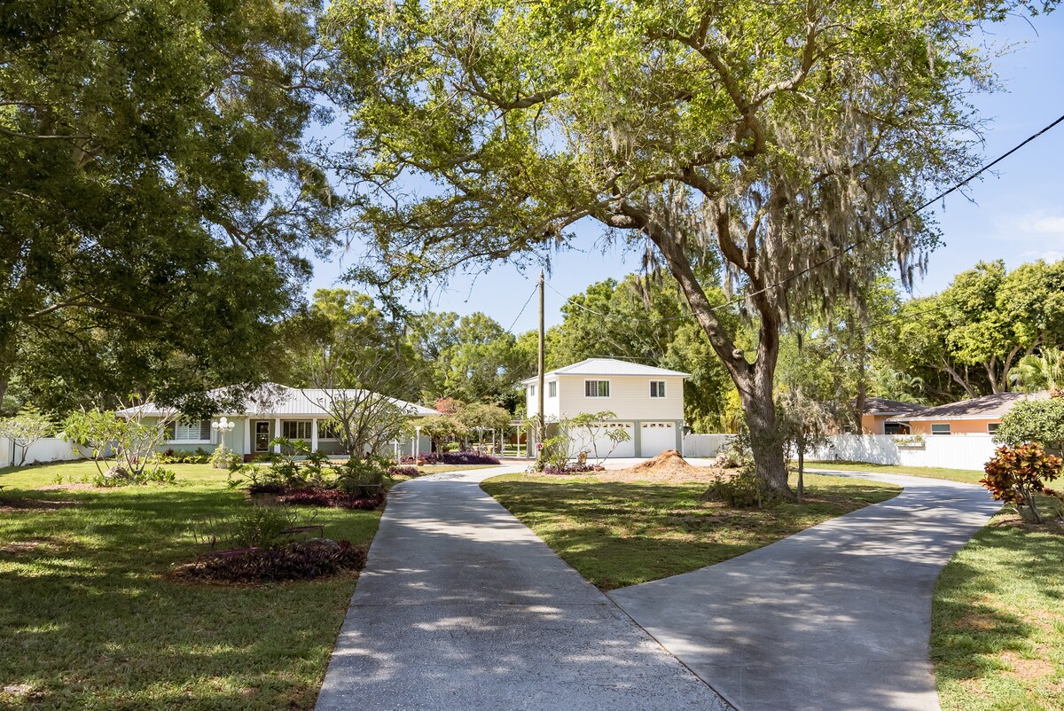 A winding driveway leads through a landscaped yard adorned with trees and shrubs. A main house and separate structure are visible in the background, both nestled under a canopy of greenery, creating a peaceful neighborhood setting.