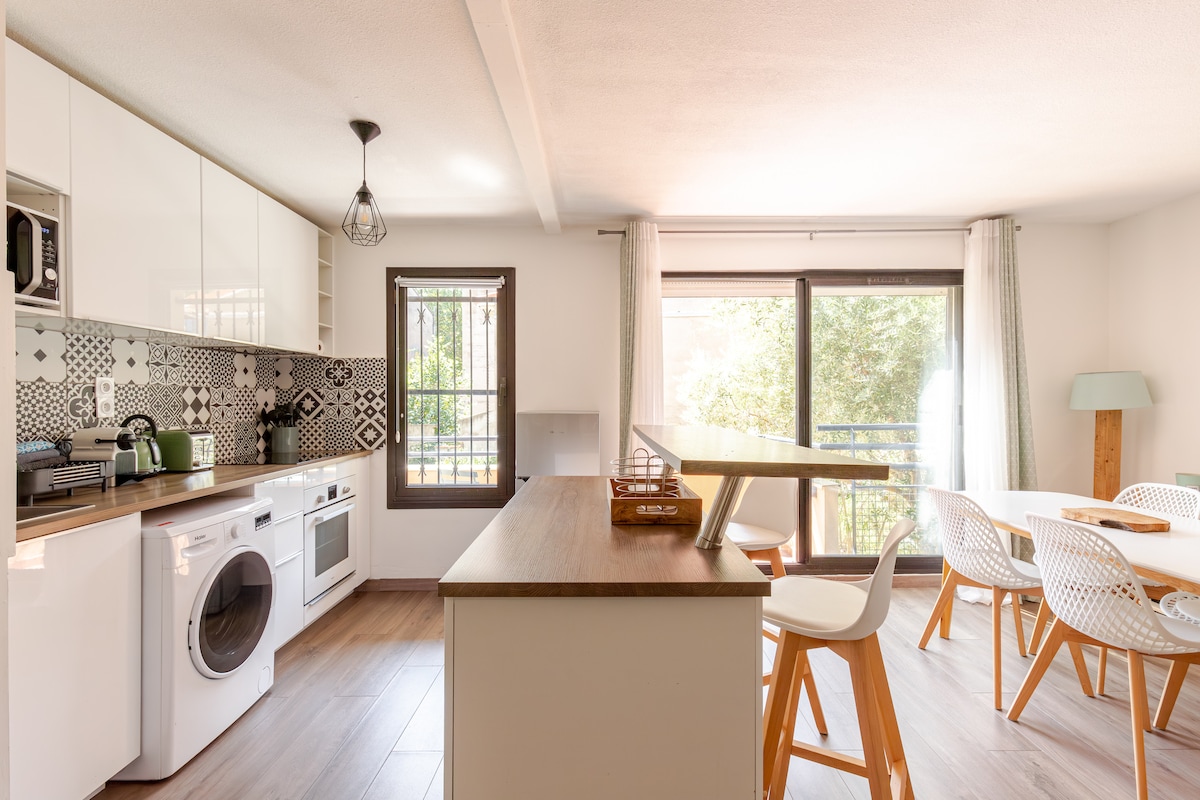 The open kitchen features modern appliances, including a washing machine and oven. A wooden countertop divides the kitchen from the living area, where a dining table and chairs are situated. Large windows allow natural light to fill the space, with greenery visible outside.