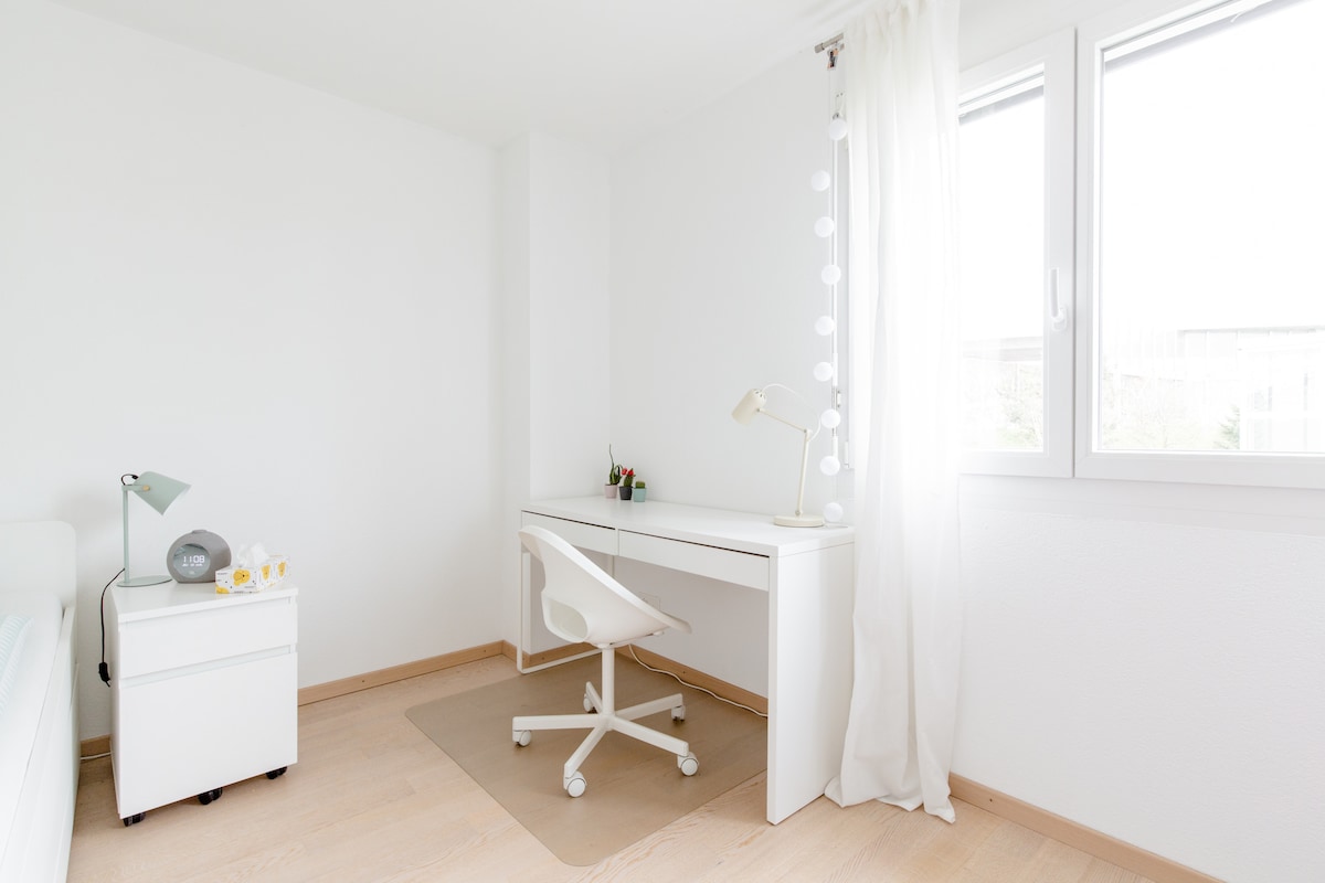 A bright and airy room is showcased with a spacious desk and a comfortable chair. A small nightstand is positioned beside the bed, and natural light filters through sheer curtains from the window, enhancing the minimalist decor.