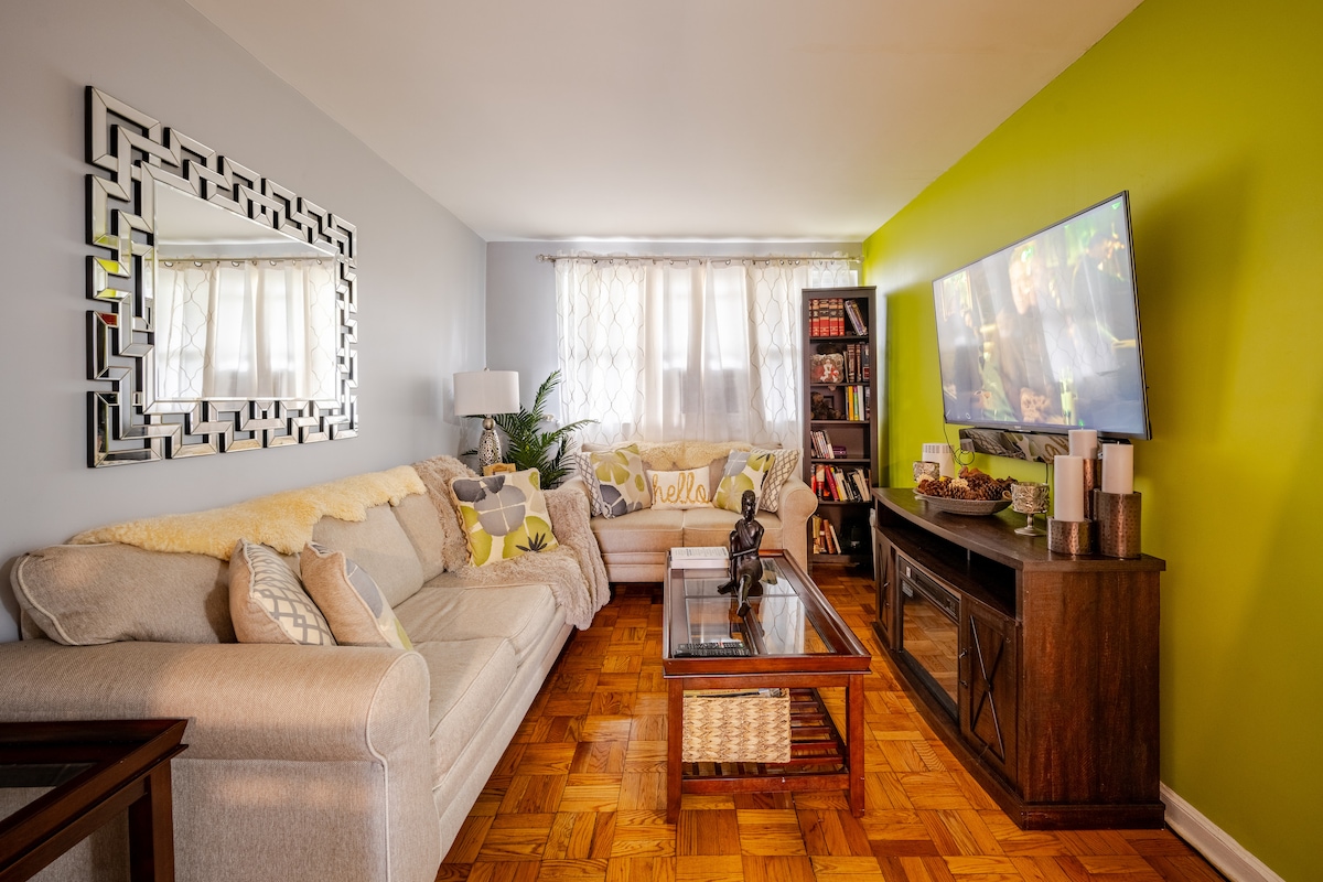 The living room showcases a spacious sectional sofa adorned with decorative pillows and a coffee table with a glass top. A wall-mounted TV is visible, accompanied by a striking green accent wall and a large mirror that reflects natural light from the windows.