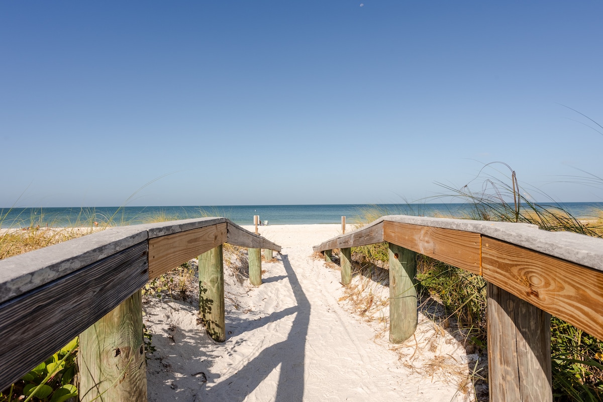 A wooden walkway leads to a serene beach, framed by soft sand and lush greenery. The tranquil ocean and clear skies are visible in the distance, inviting relaxation and outdoor activities. The scene captures a peaceful journey from the pathway to the shoreline.