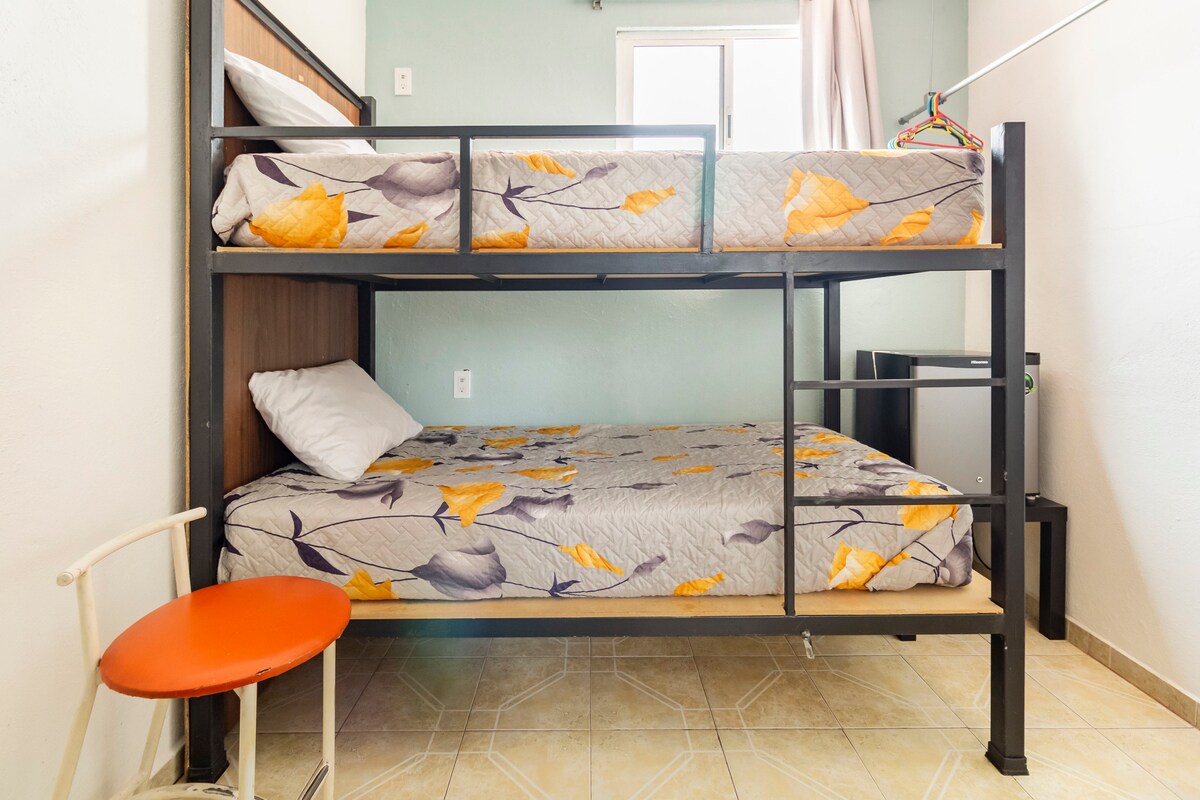 A compact sleeping area features a sturdy bunk bed with floral-patterned bedding, accommodating multiple guests. A small round table and a chair are positioned nearby. Natural light enters through a window, illuminating the space with a soft glow.
