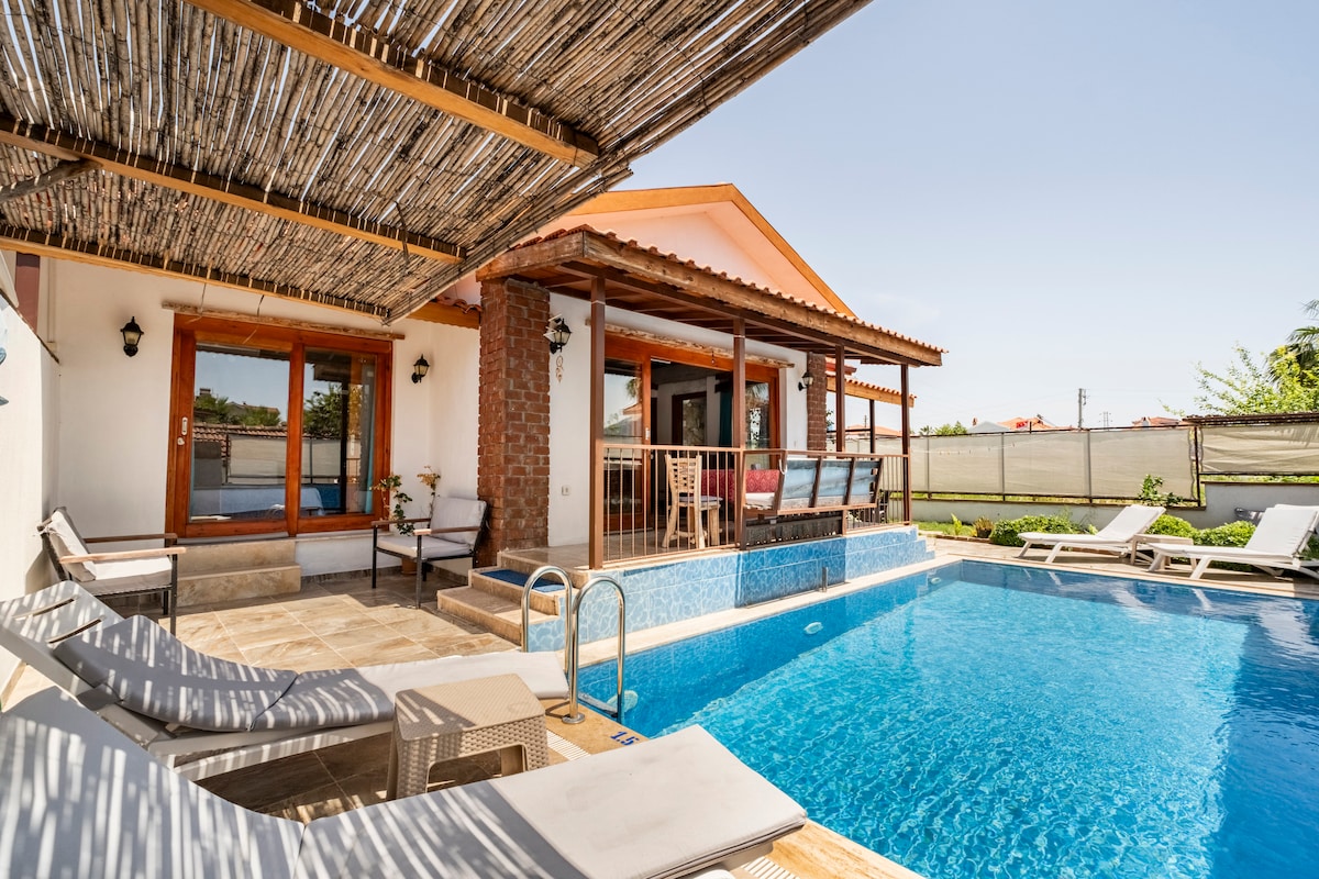A private pool area features a clear blue pool surrounded by sun loungers on a wooden deck. A shaded terrace with wooden beams provides shelter and includes seating. Large glass doors lead into an indoor living space, with greenery visible in the background.