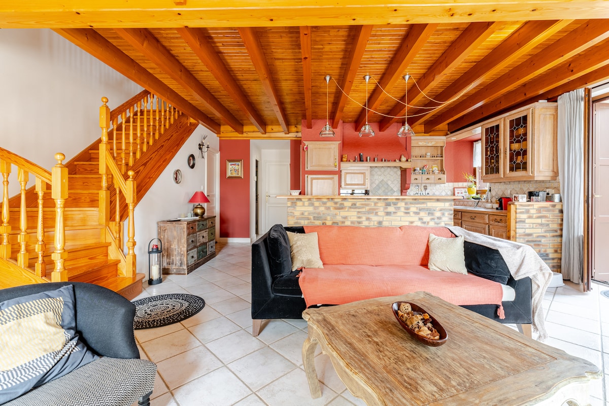 A welcoming living area is highlighted by exposed wooden beams above. Two comfortable sofas are arranged around a wooden coffee table, with a staircase in the background leading to upper levels. The open kitchen features wooden cabinetry, and warm tones create an inviting atmosphere.