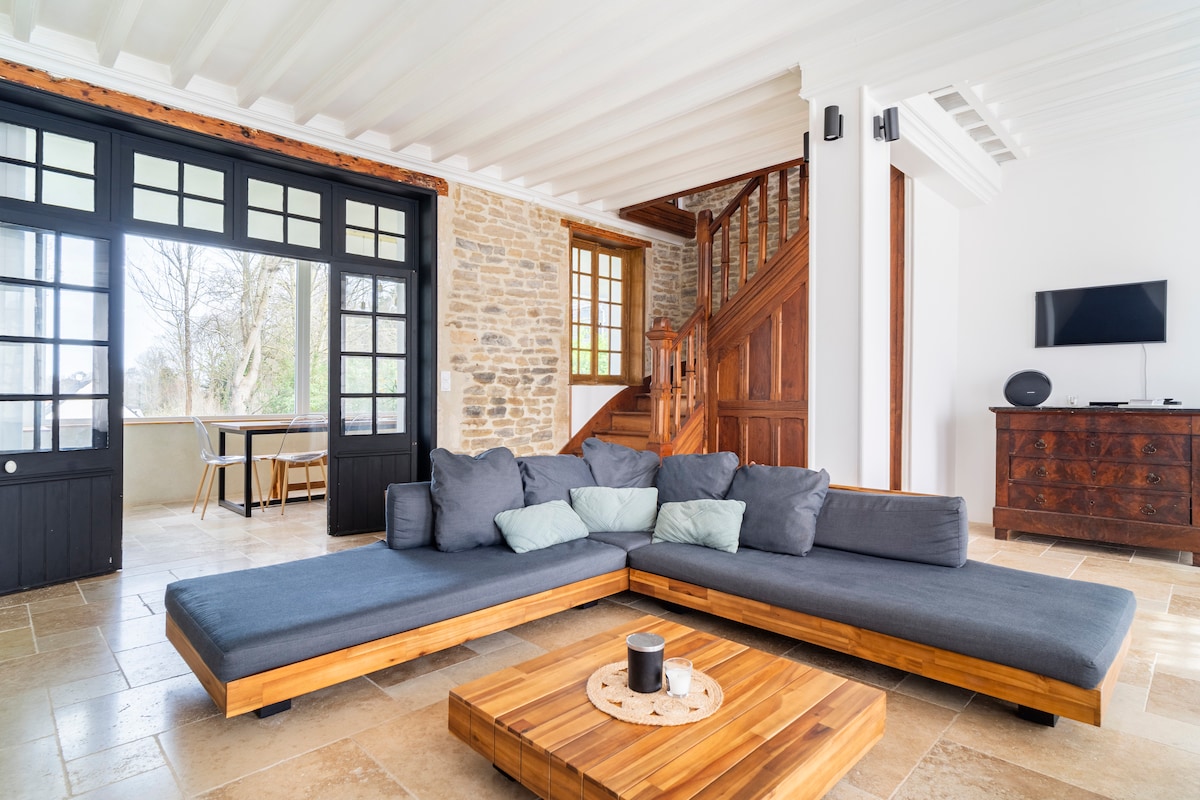 A spacious living area features a large sectional seating arrangement in gray fabric with wooden accents. A coffee table sits at its center, accompanied by decorative cushions. Natural light fills the room through large windows, while a stylish staircase and a dark wooden sideboard add character.
