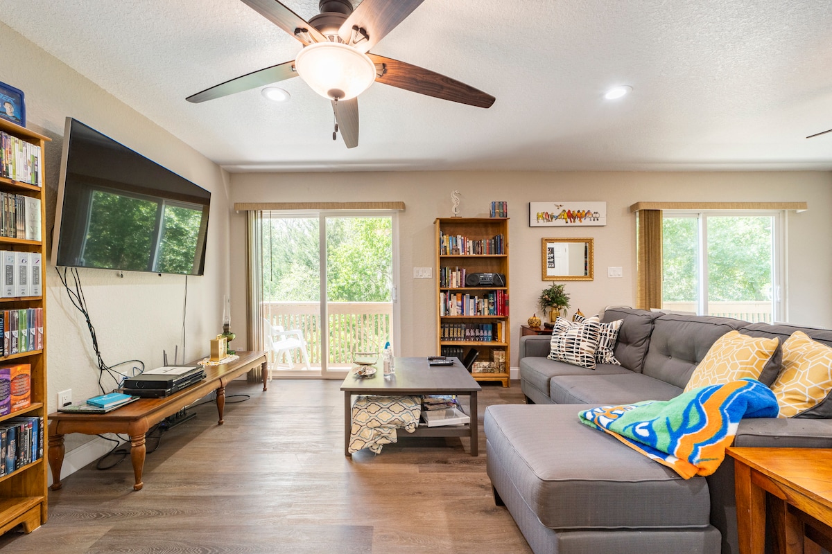 A spacious living area is illuminated by natural light from large windows. A comfortable sectional sofa is accompanied by a coffee table, and a fan hangs from the ceiling. A TV is mounted on the wall, with shelves displaying books and decorative items.