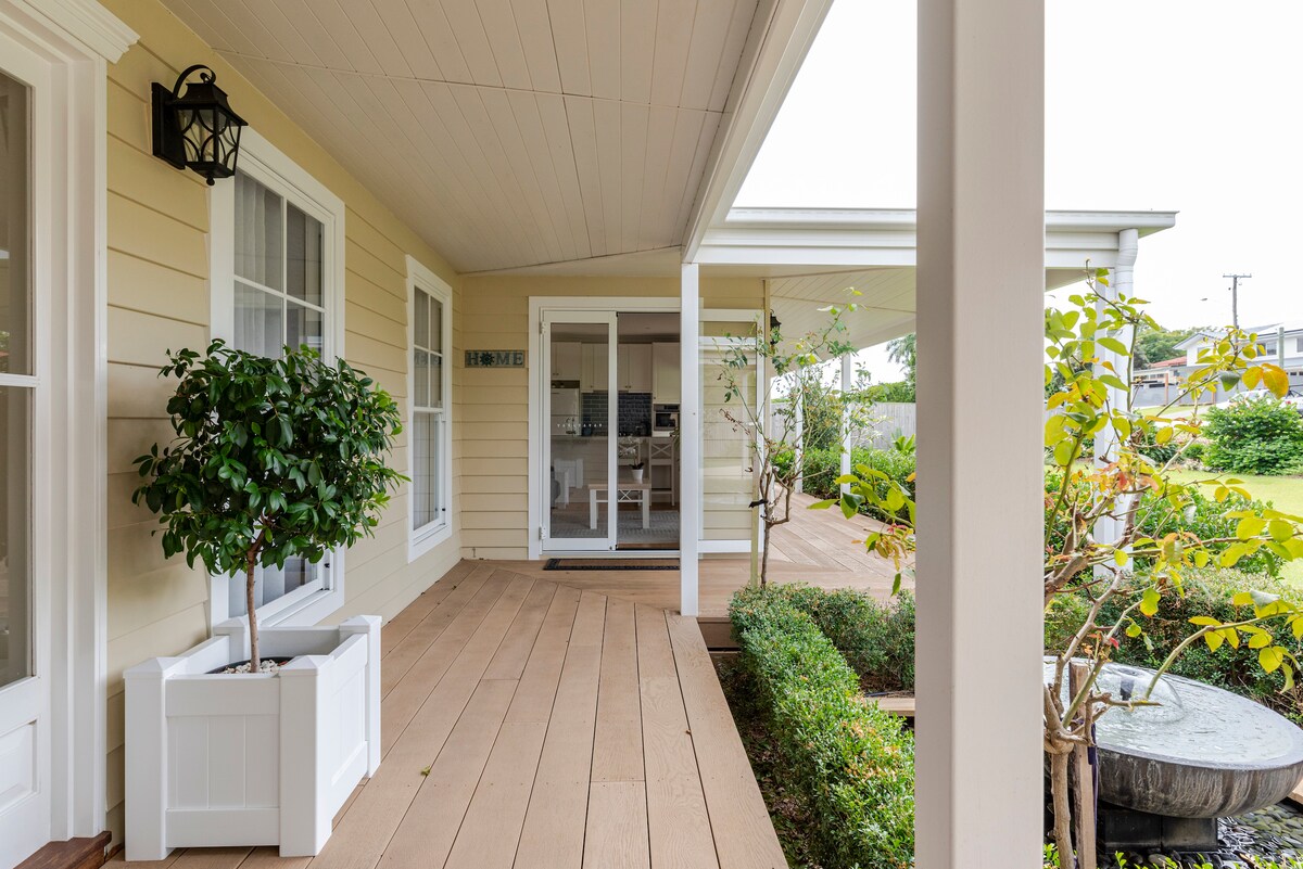 A covered verandah features wooden decking, with potted greenery adding a natural touch. Glass doors provide a view into the kitchen area, while manicured hedges and a rock water feature create a serene outdoor environment.
