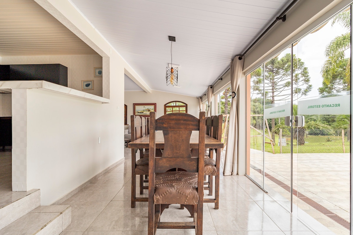 A spacious dining area features a large wooden table surrounded by several chairs. Floor-to-ceiling glass doors provide views of the outdoor space, which is filled with greenery. Neutral colors create a calm environment, with natural light streaming into the room.