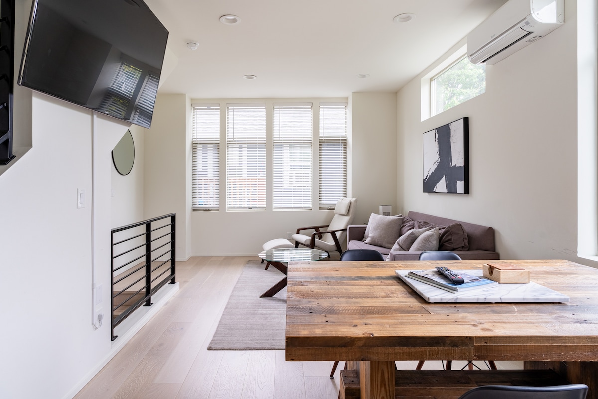 A welcoming living area features a comfortable sofa and an armchair arranged around a wooden coffee table. A television is mounted on the wall opposite a large window with blinds. Natural light fills the space, highlighting the wooden dining table adorned with magazines.