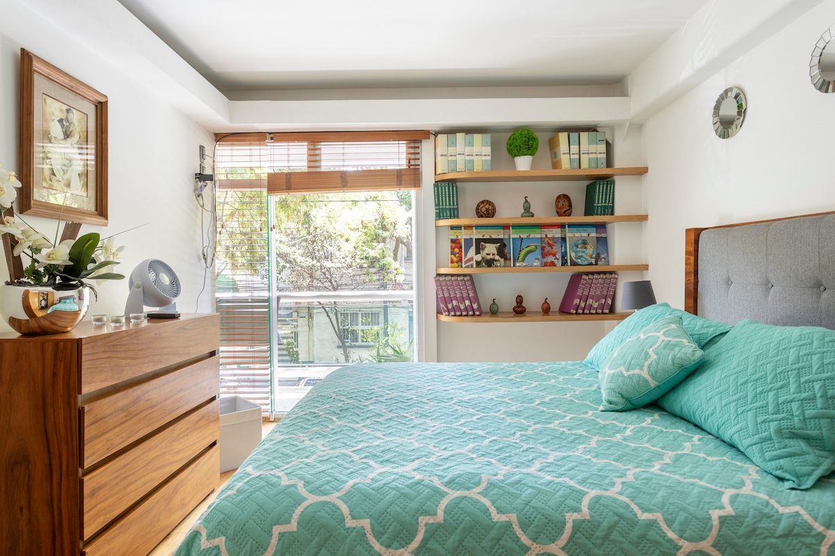 A cozy bedroom features a spacious bed dressed in a teal quilt, complemented by wooden furniture. Shelves adorned with books and decorative items create a welcoming atmosphere, while large windows allow natural light and offer a view of greenery.