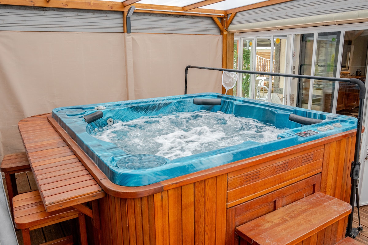 An inviting spa area is seen within a sheltered structure, featuring a blue hot tub surrounded by wooden panels. Adjacent to the tub, a wooden bar with stools provides a space for relaxation. Natural light filters through the translucent roofing.
