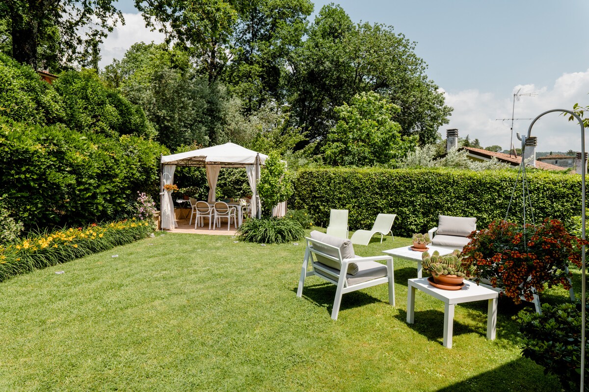 A neatly maintained garden is visible, featuring a spacious green lawn surrounded by lush greenery. White lounge chairs and tables are arranged neatly, along with a shaded gazebo providing additional seating. Flowers bloom along the edge, adding color to the serene outdoor space.