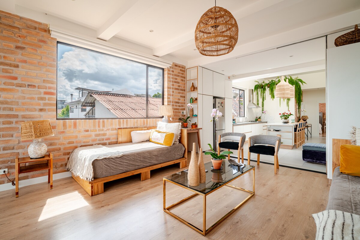 A bright living space is highlighted by a large window showcasing city views. Natural materials, including a wooden bed and brick walls, accentuate the decor. A glass coffee table is surrounded by various textures, and a potted plant adds a touch of greenery to the room.