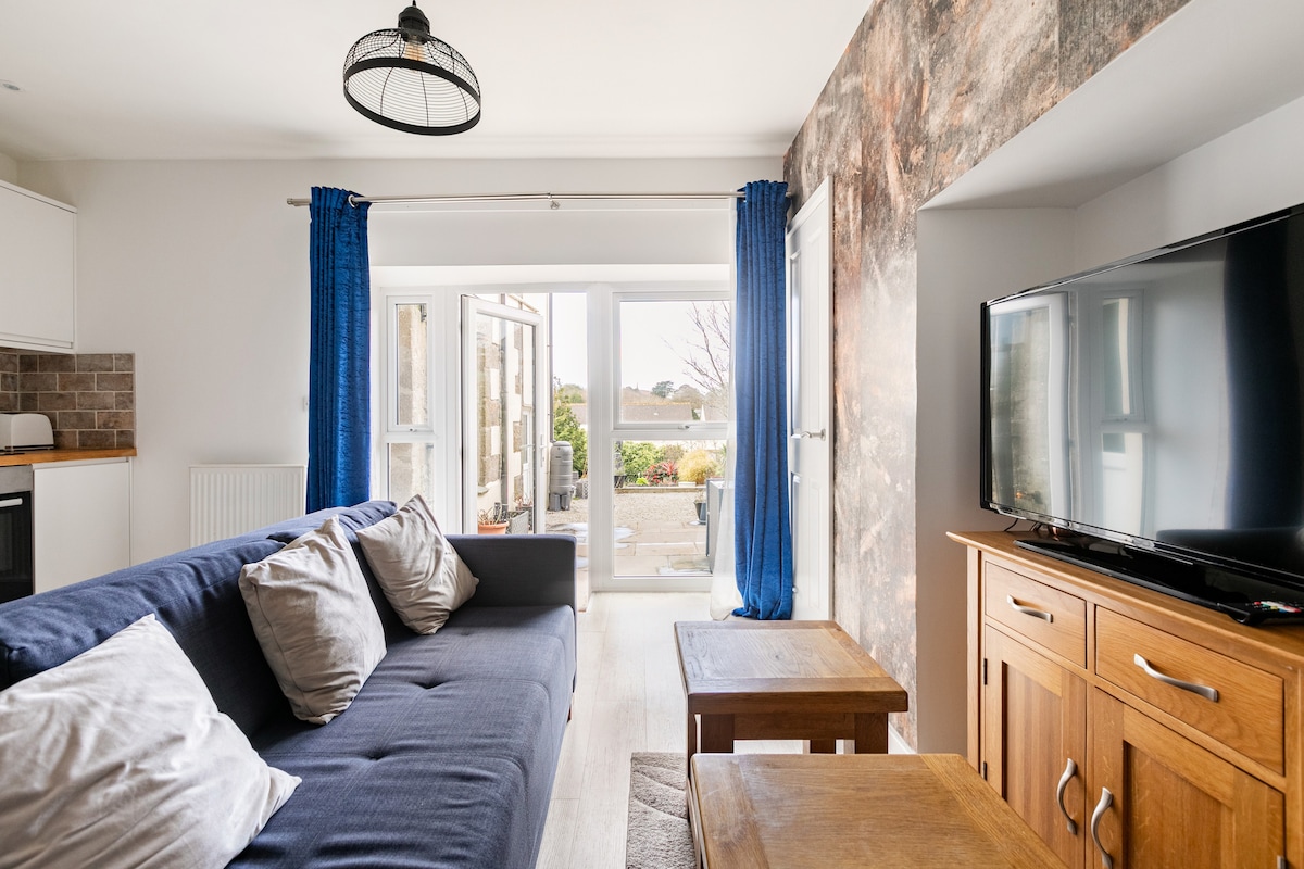 The lounge area features a comfortable sofa adorned with cushions, positioned in front of a flat screen television. Large glass doors open to a private courtyard garden, complemented by blue curtains that frame the space. A wooden cabinet adds warmth to the modern décor.