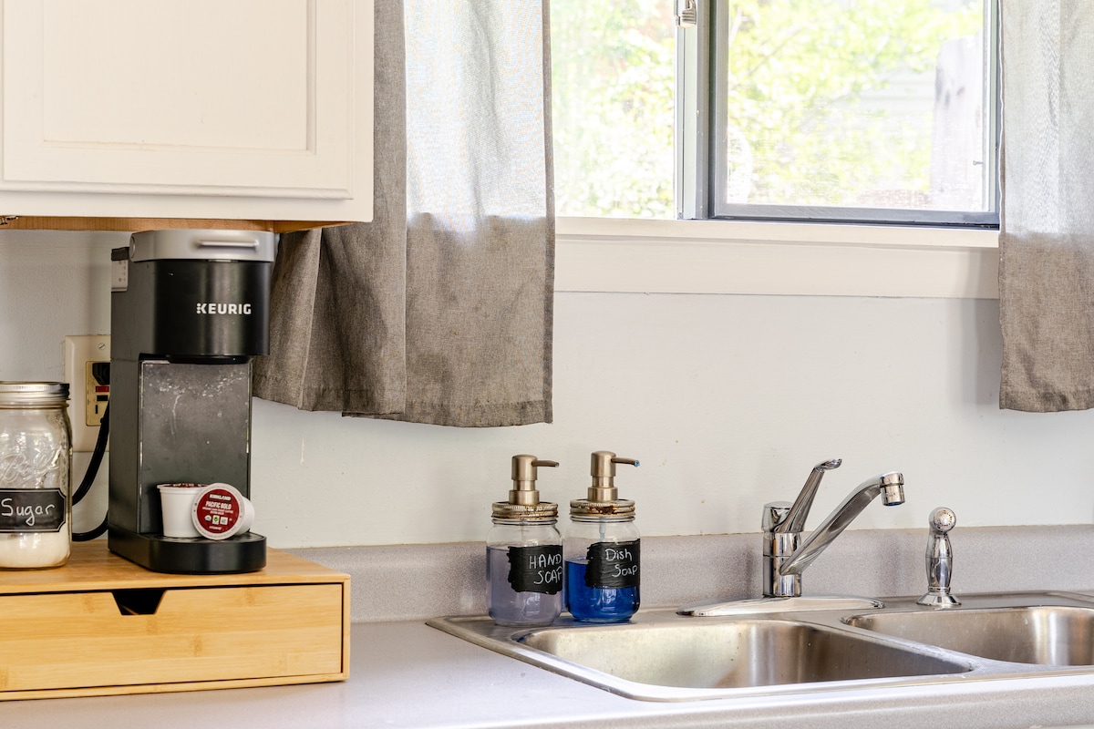 A kitchen counter displays a coffee maker alongside labeled glass containers for sugar and dish soap. Natural light filters in through a nearby window, revealing a neutral-toned wall and a clean, organized sink area.